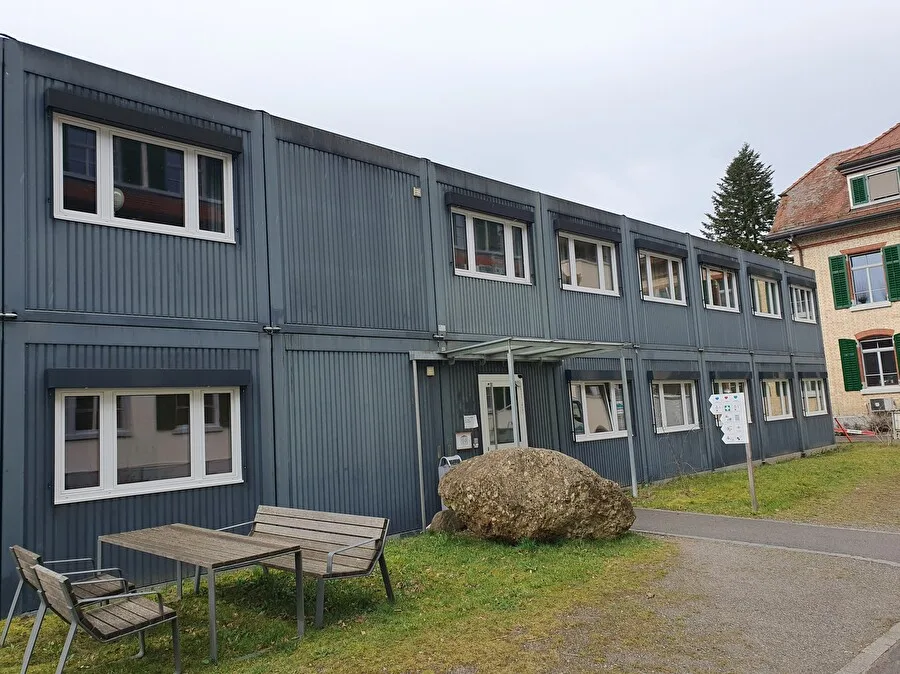Blaue Wohncontainer stehen an einem Weg mit einem Wiesensaum. Im Vordergrund ist eine Sitzgruppe mit Bänken und einem Tisch aus Holz.